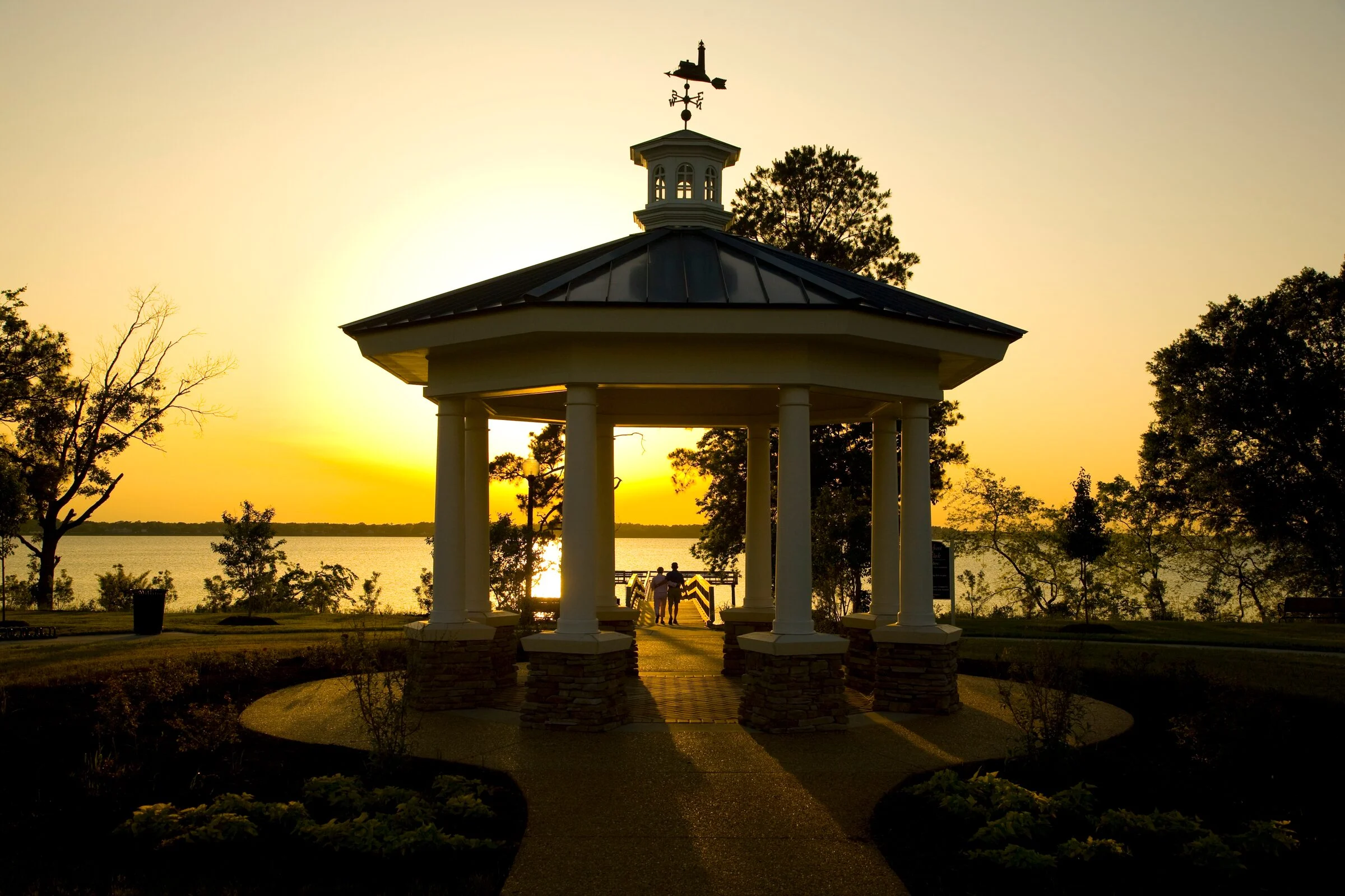 The River Park gazebo at sunset, with a couple on the pier beyond it and the river on the horizon