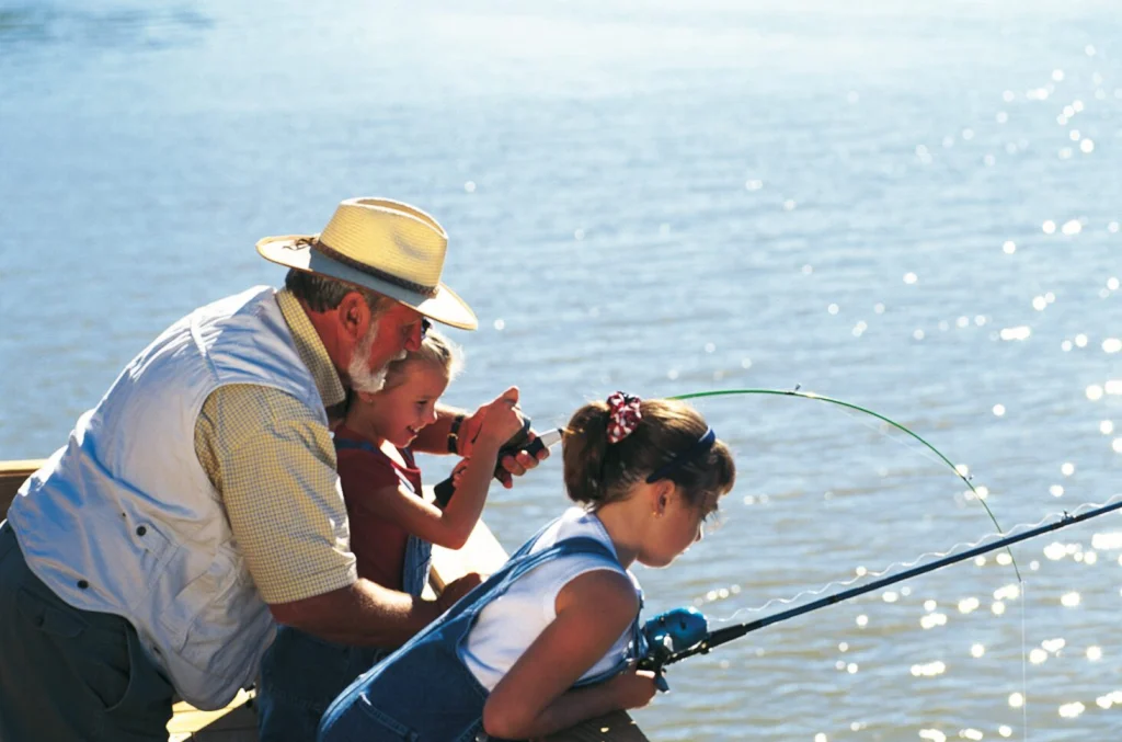 A grandfather in a straw hat fishing from a pier with two young girls
